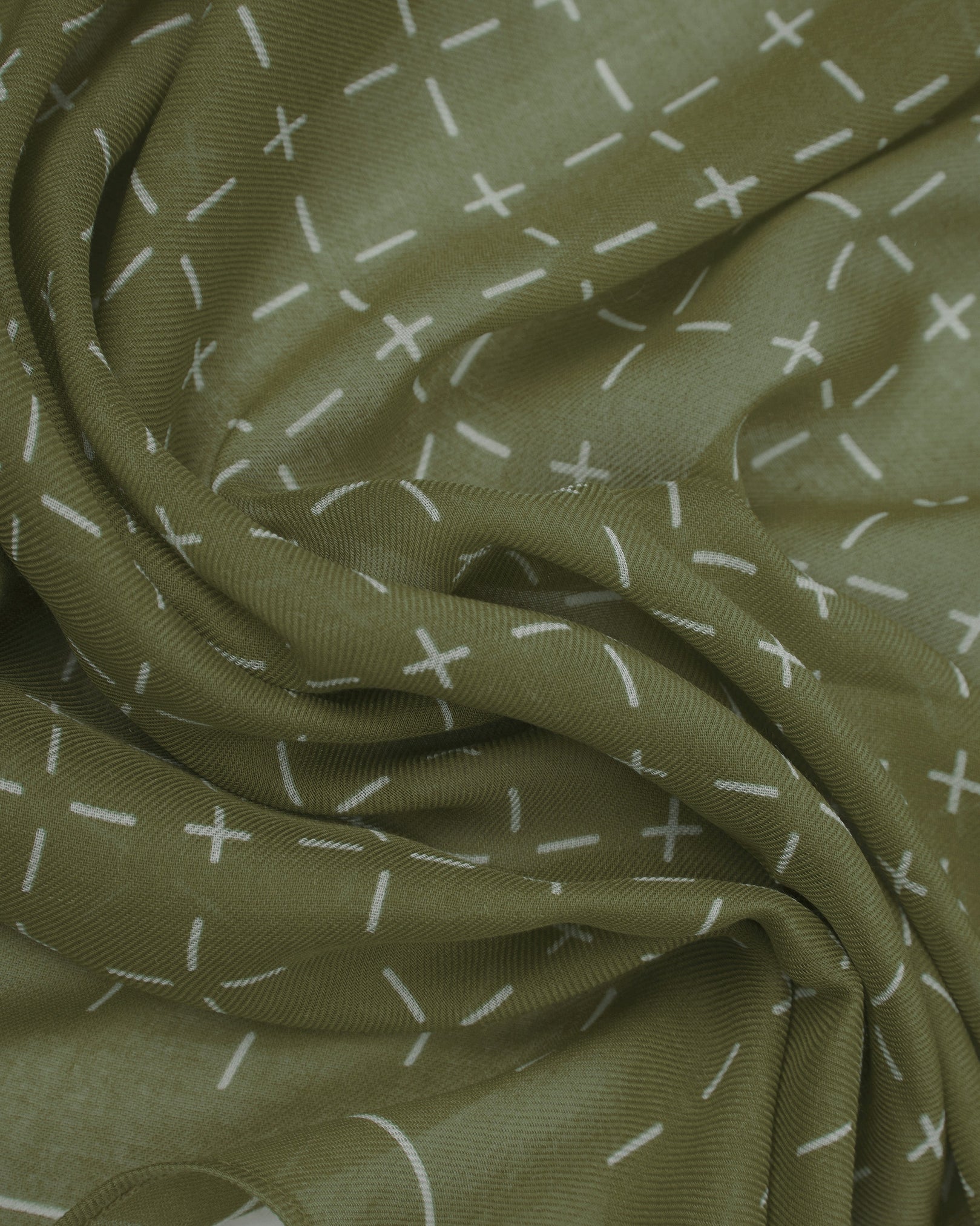A ruffled close-up of the 'Crosses in Olive' modal and wool bandana from SOHO Scarves, highlighting the soft modal and wool blend fabric with a repeating geometric grid pattern of dashes and crosses in muted olive and contrasting tones.