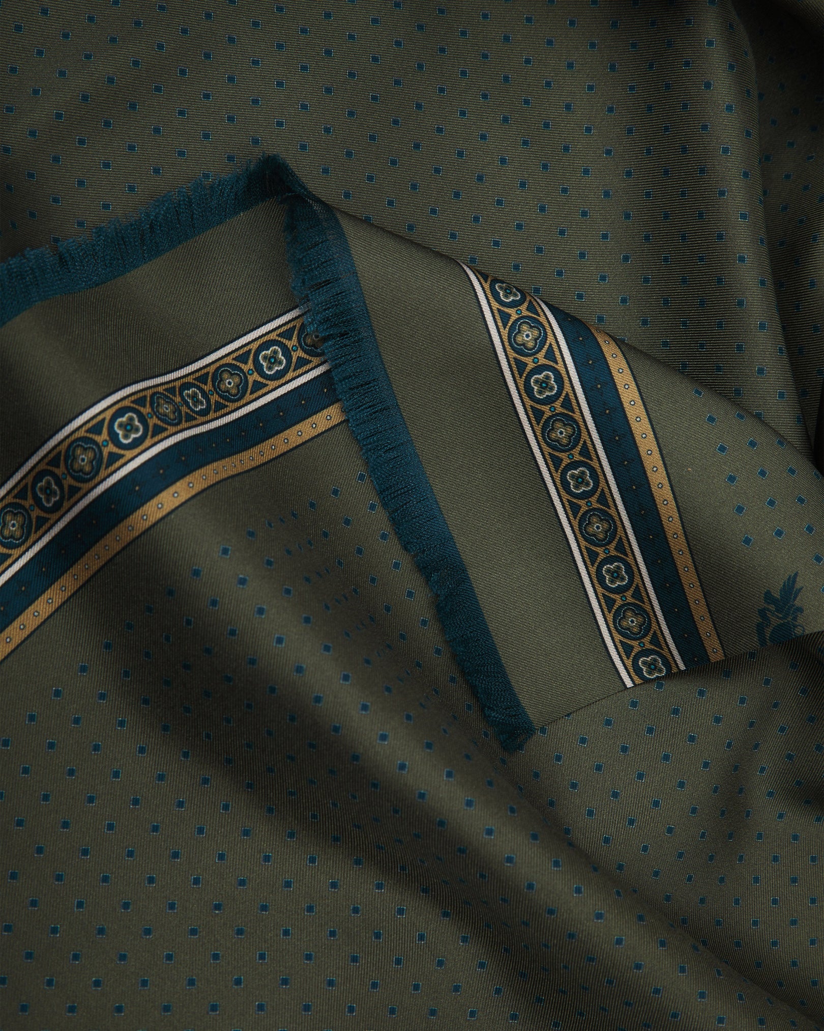 A ruffled close-up of the 'Chiswick' silk scarf from SOHO Scarves, showcasing the luxurious drape of 16 momme silk twill on a warm military olive ground with a subtle micro-dot pattern, finished with an elegant ornamental border.