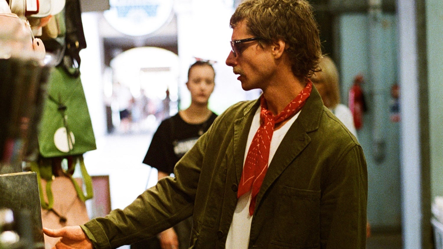 A man wearing a dark olive green blazer, white trousers, and a red patterned neck scarf stands in a market alley, gesturing toward merchandise on a display stand.
