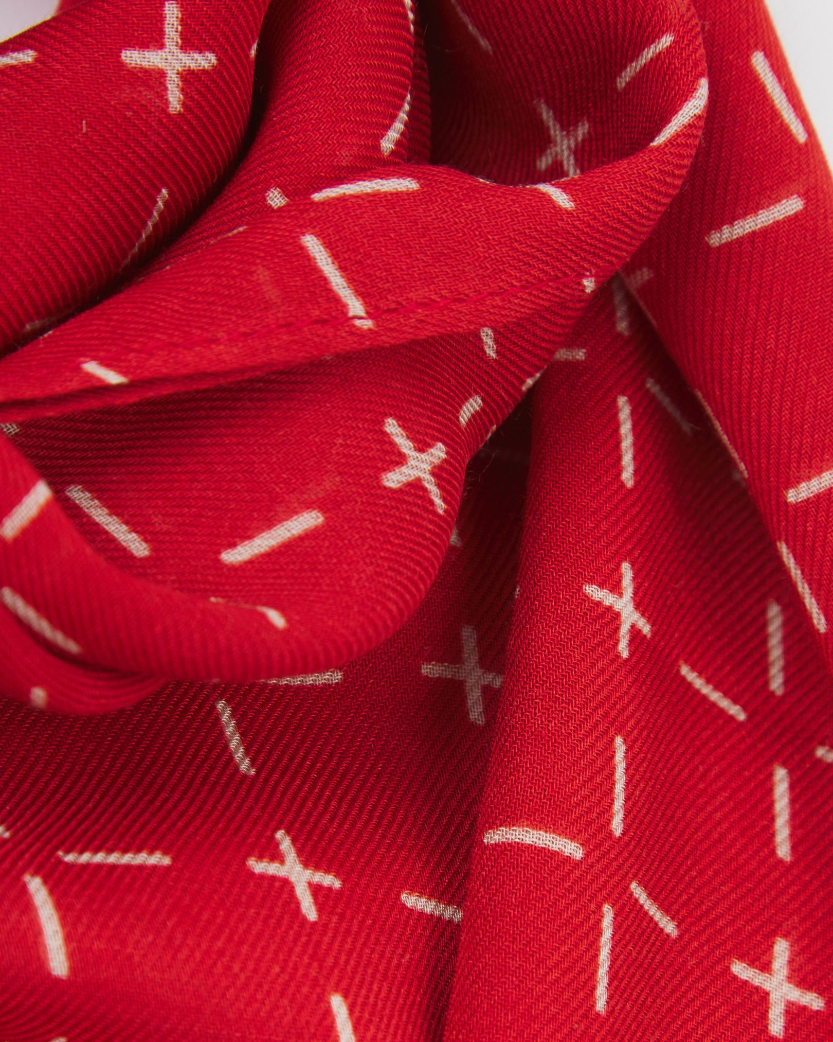 A ruffled close-up of the 'Crosses' red bohemian wide scarf, showing the soft modal and wool blend fabric with a repeating geometric grid of white dashes and crosses on a bright red background.
