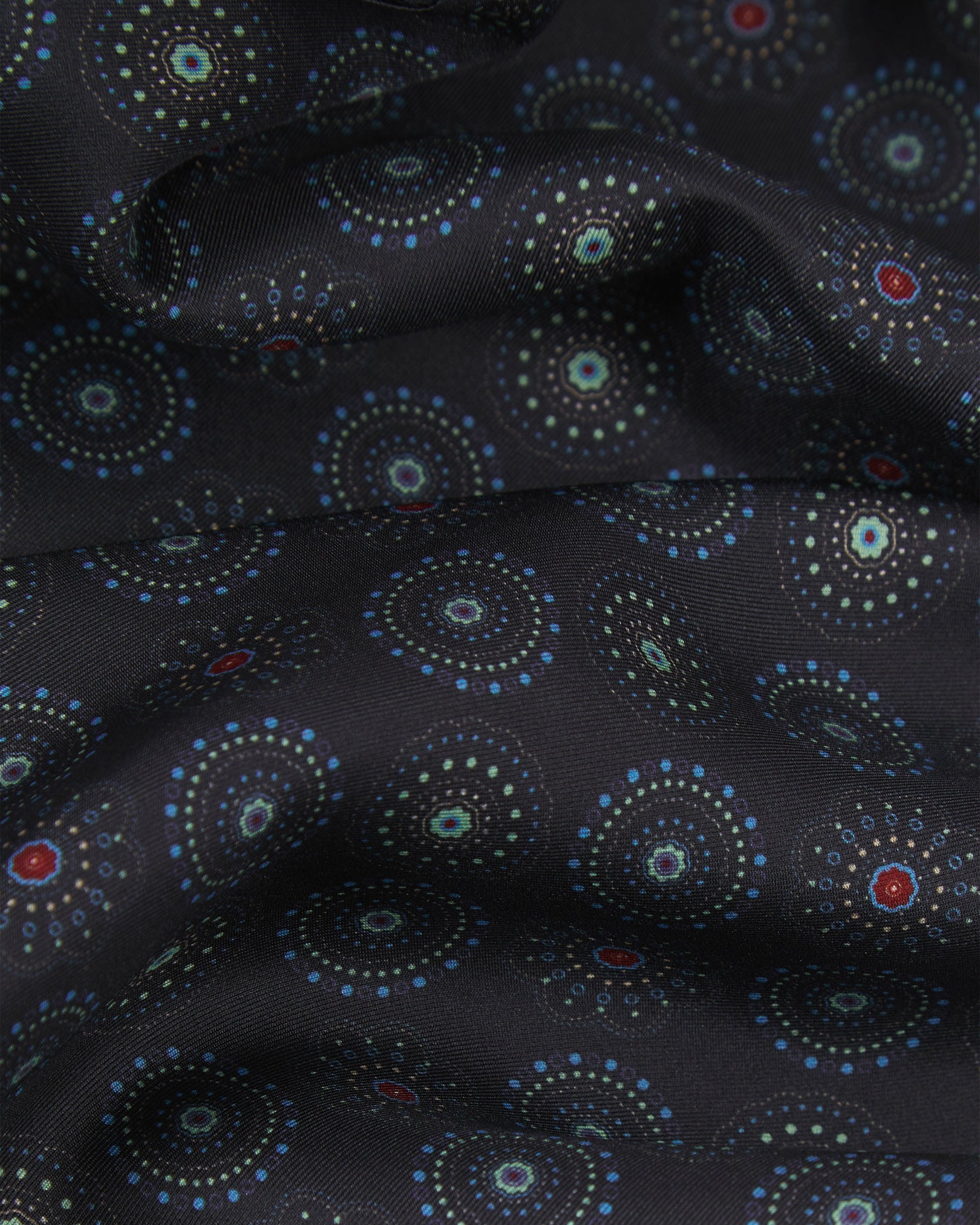 A ruffled close-up of the Seattle 'Gardenia' wool-backed silk dress scarf, showing the soft silk fabric with intricate circular motifs in blue and mint green on a black background with subtle red accents.
