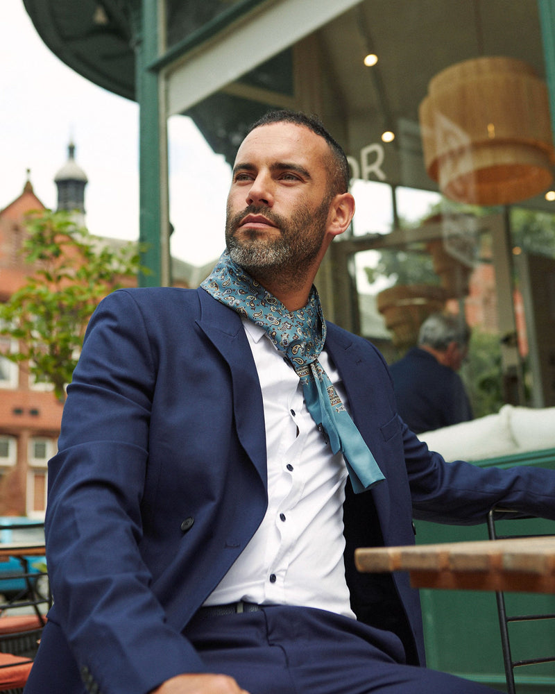 Outdoor shot of model seated wearing peacock blue 'The Banff' paisley polyester scarf paired with white shirt and casual navy suit.