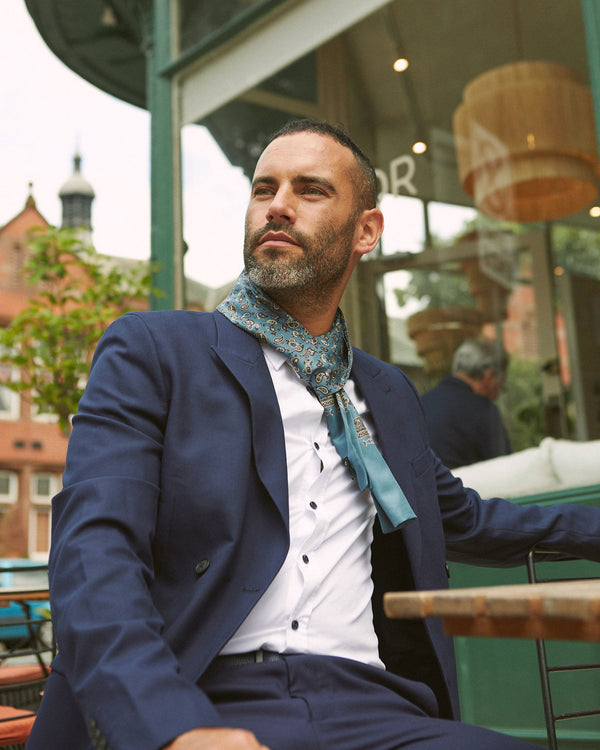 Outdoor shot of model seated wearing peacock blue 'The Banff' paisley pure silk scarf paired with white shirt and casual navy suit.