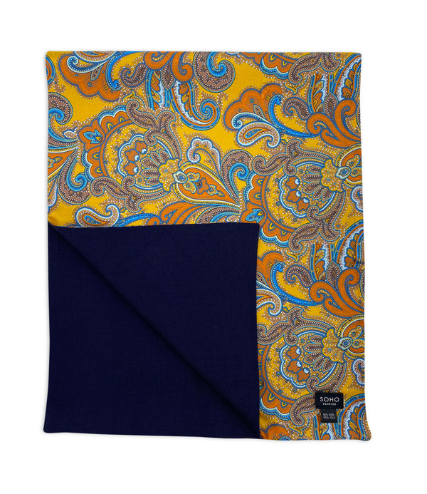 'The Carnaby' wide dress scarf arranged in a square shape clearly showing the orange, violet and powder blue paisley patterns set on a gold-yellow ground. The bottom-left quadrant is folded back to reveal the fine woollen lining underneath the pure silk exterior.