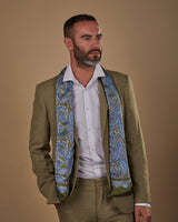 Slightly angled to his left wearing an Abraham wool-silk scarf in blue paisley with olive green ground. Scarf draped loosely over neck and shoulders.