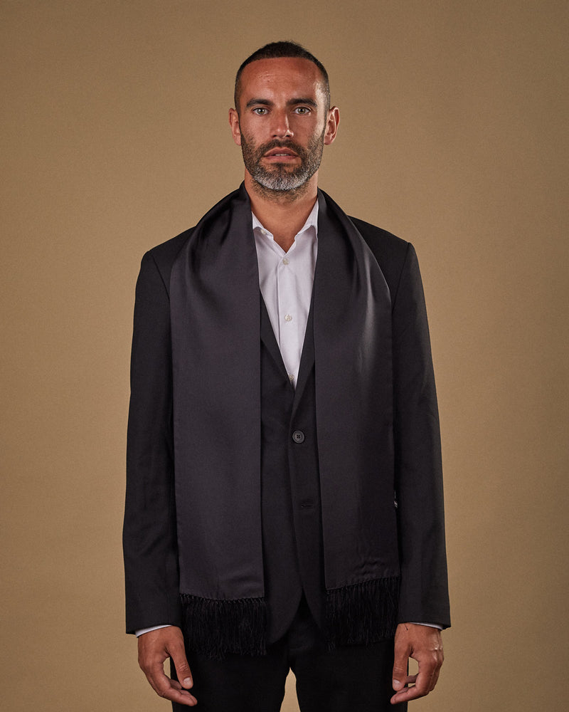 Portrait view of male model looking straight ahead, wearing 'The Air Black' silk aviator scarf. Draped loosely around shoulders and neck and paired with a smart, dark suit.