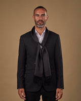 Portrait view of male model wearing 'The Air Black' silk aviator scarf. Tied loosely around neck in a simple knot and paired with a smart, dark suit.