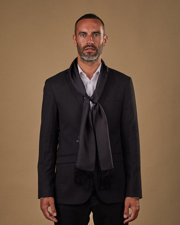 Portrait view of male model wearing 'The Air Black' silk aviator scarf. Tied loosely around neck in a simple knot and paired with a smart, dark suit.