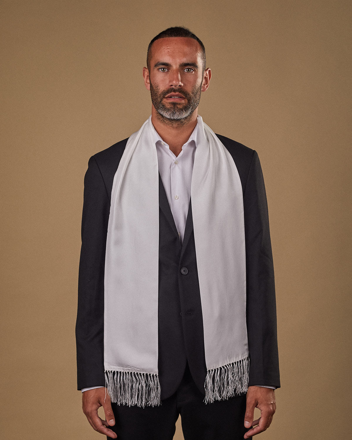 Portrait view of male model looking straight ahead, wearing 'The Air White' silk aviator scarf. Draped loosely around shoulders and neck and paired with a smart, dark suit.
