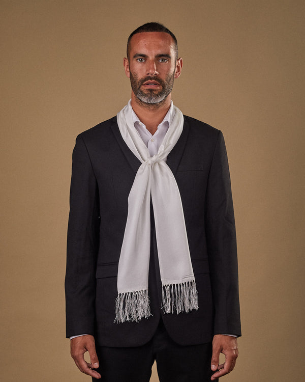 Portrait view of male model wearing 'The Air White' silk aviator scarf. Tied loosely around neck in a simple knot and paired with a smart, dark suit.