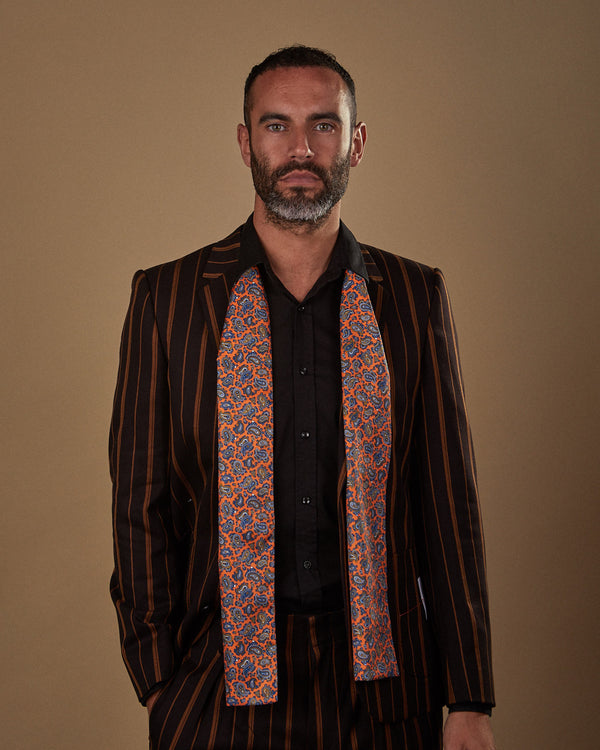 Model facing ahead with hand in pocket, wearing the 'Asakusa' polyester scarf featuring small blue paisley patterns with an orange ground. Paired with copper striped black suit and dark shirt.