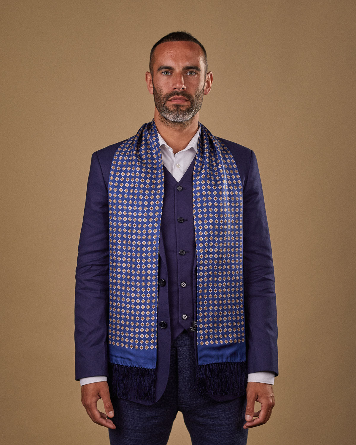 Portrait view of male model looking straight ahead, wearing 'The Auburn' silk aviator scarf. Draped loosely around shoulders and neck and paired with a smart, midnight-blue suit.