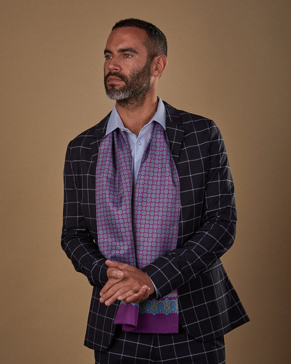 Three quarters view looking to his right wearing 'The Beak' purple and blue geometric patterned polyester scarf draped around neck and paired with pale blue shirt and dark chequered suit.