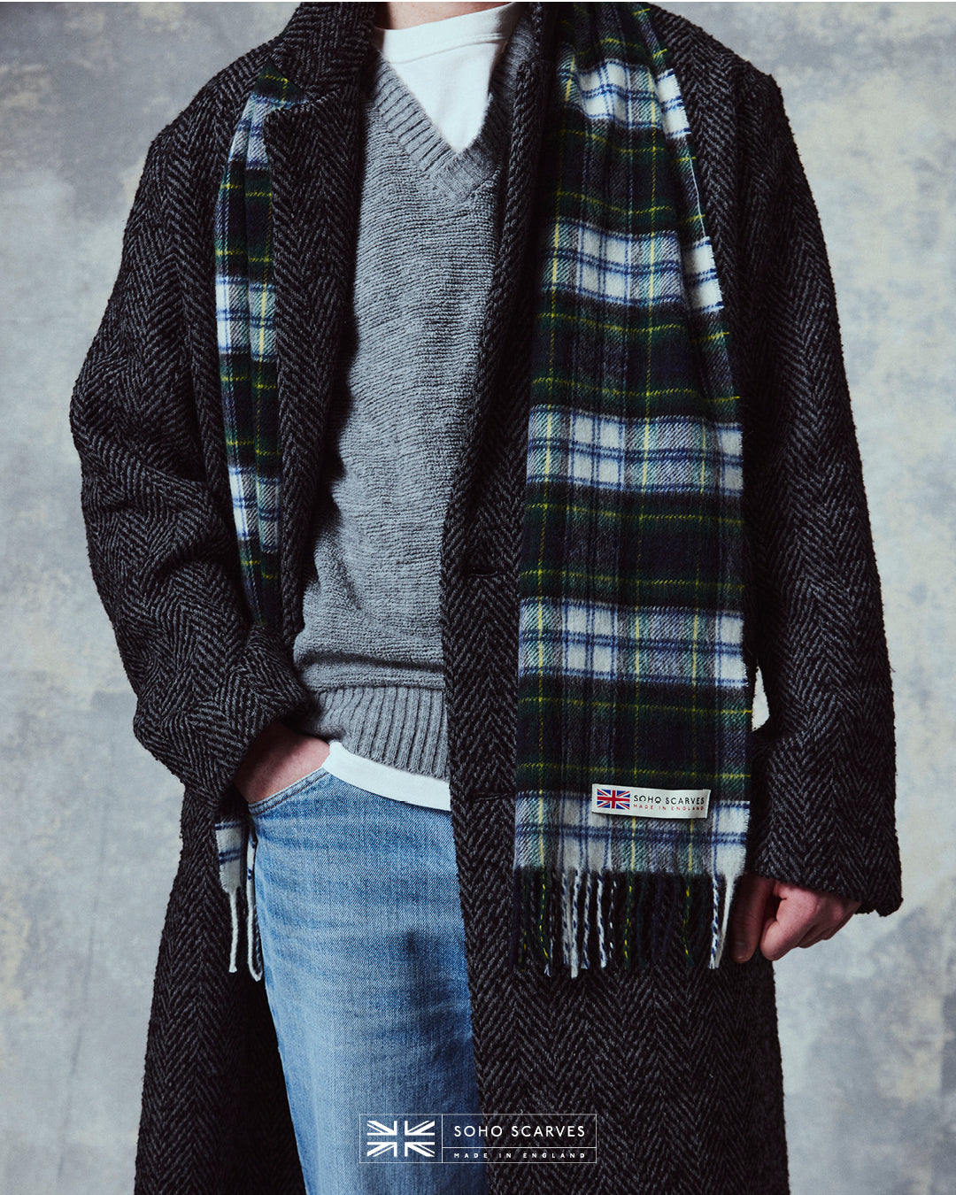 Bottom-half view of man wearing the Yorkshire-made, 'Highlands Tartan in Grey' wool scarf, with focus on the 'Soho scarves, Made in England' label and matching fringes.