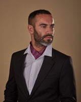 Model looking to his left, wearing red and blue geometric patterned 'Canaria' single pointed Ascot tie. Neatly tucked under white shirt and paired with charcoal jacket.