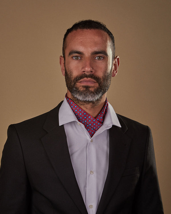 Model looking straight ahead wearing red and blue 'Canaria' single pointed Ascot tie tucked neatly under white shirt. Paired with charcoal jacket.