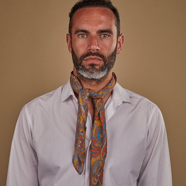 Man wearing 'The Carnaby' multicoloured paisley bandana on a golden ground. Tied snugly around neck in a simple knot and paired with a white shirt.