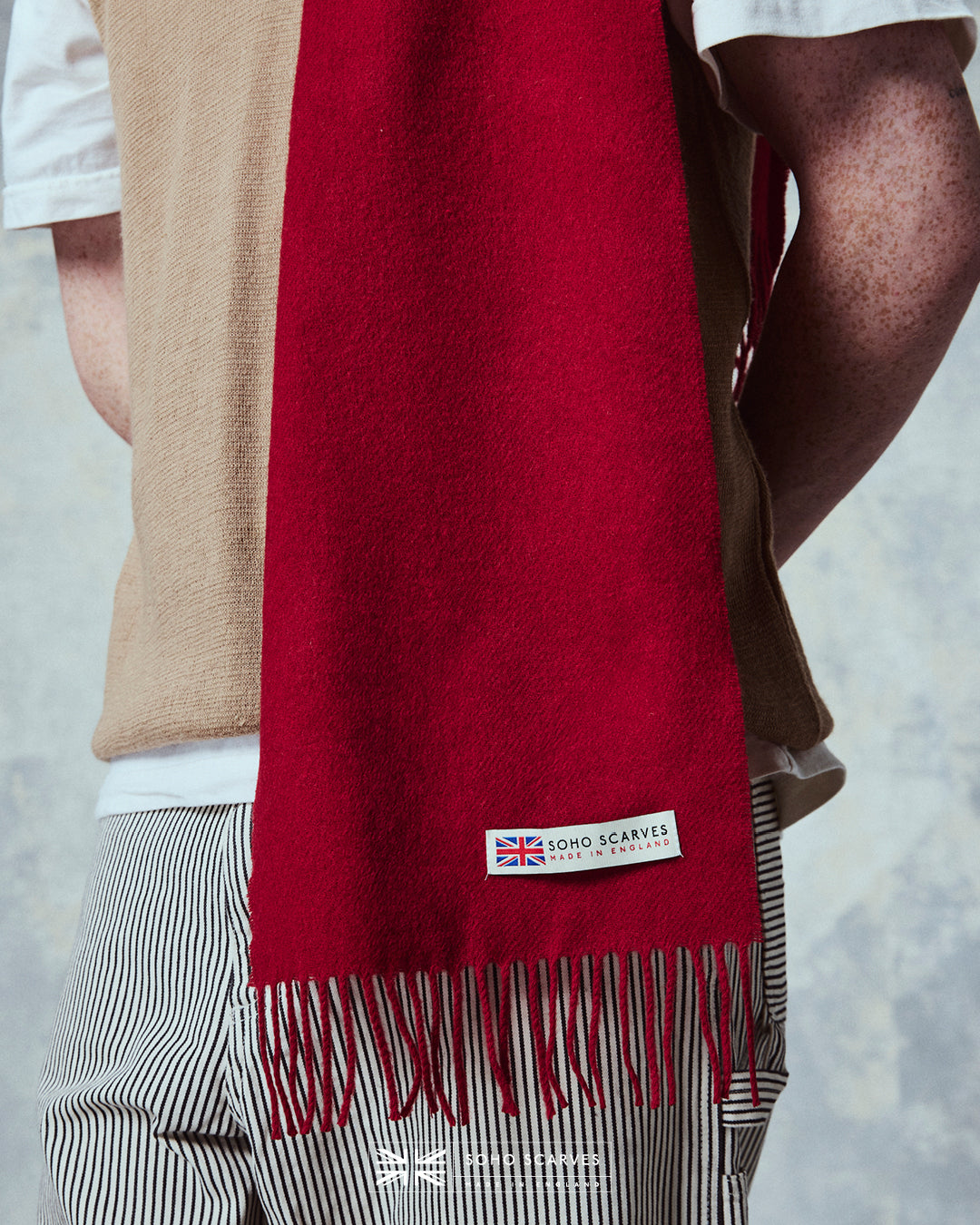Close up of man wearing the Yorkshire-made, 'Lowlands in Scarlet' wool scarf, with focus on the 'Soho scarves, Made in England' label and matching fringes.