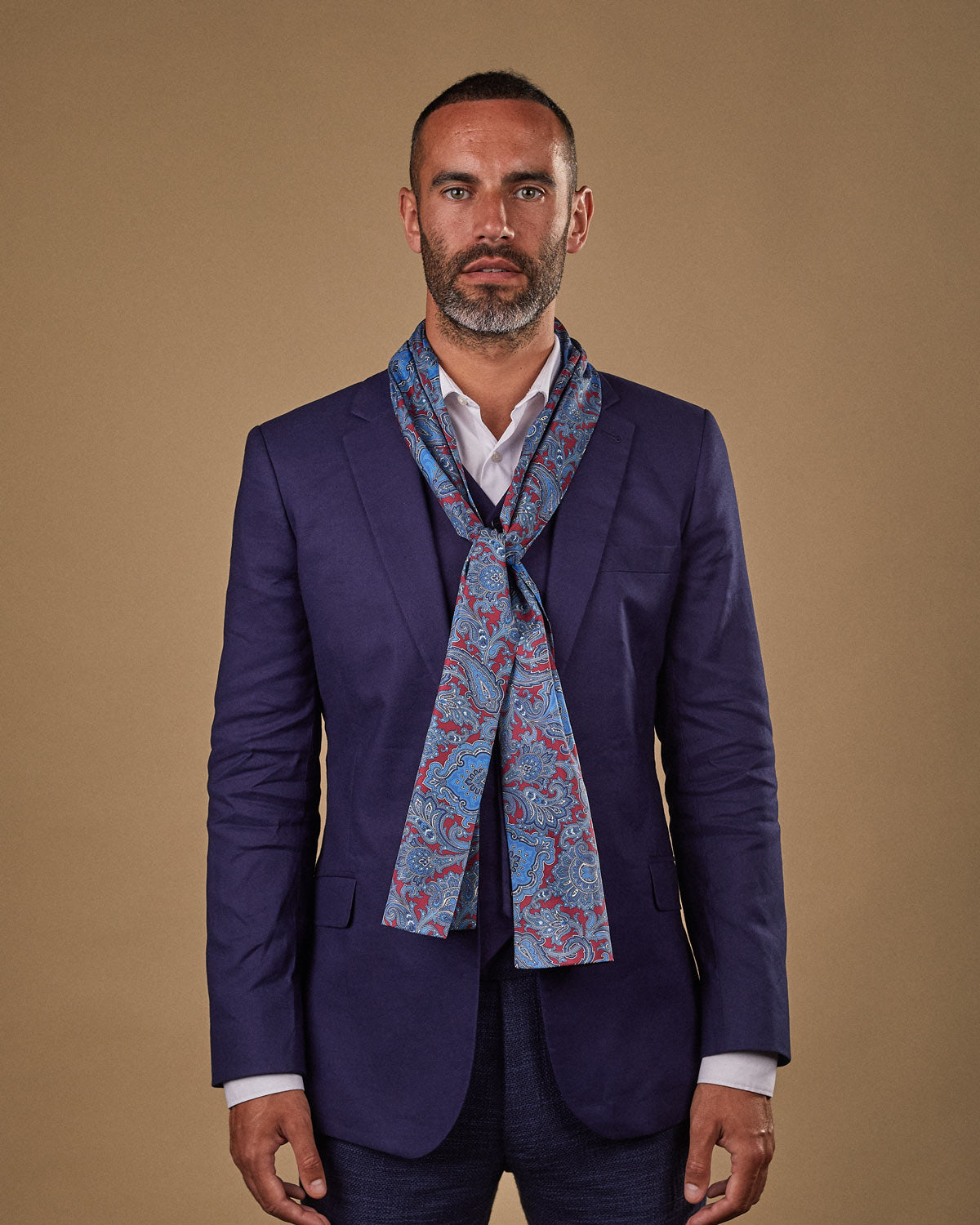 Portrait view of male model wearing 'The Dean' silk scarf. Tied loosely around neck in a simple knot and shoulders and paired with a smart, midnight-blue suit.