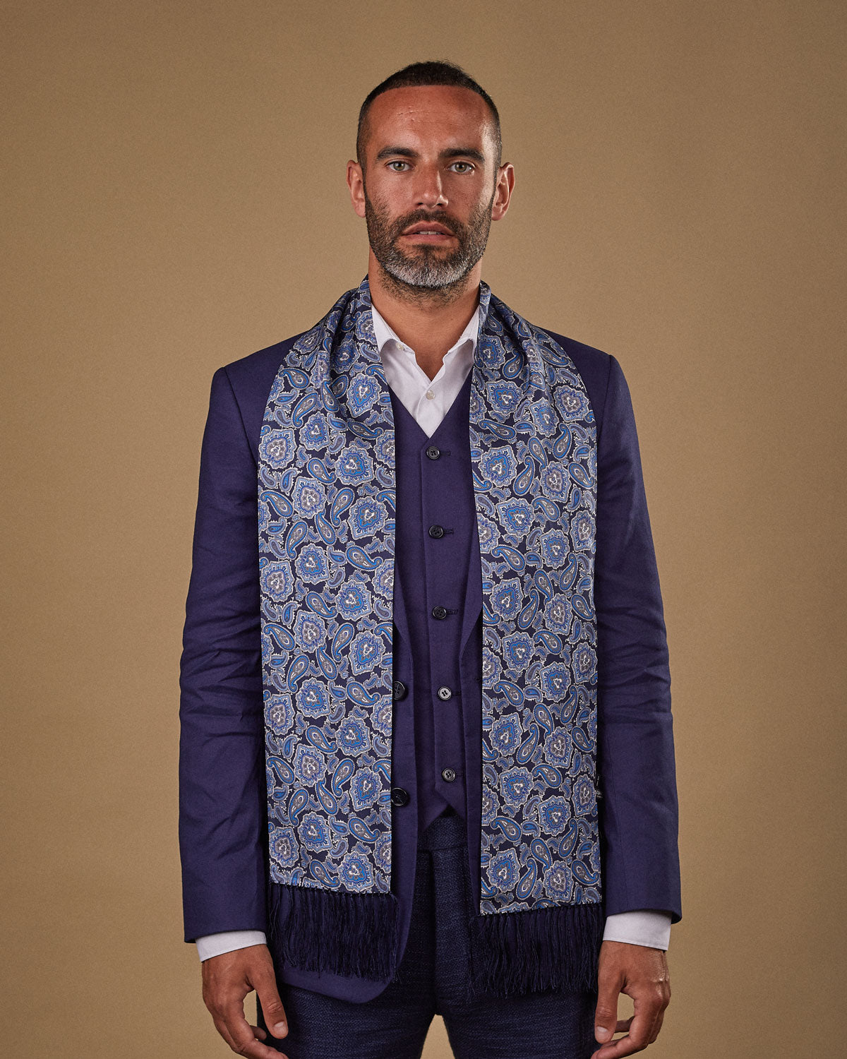 Portrait view of male model looking straight ahead, wearing 'The DeZon' silk aviator scarf. Draped loosely around shoulders and neck and paired with a smart, midnight-blue suit.