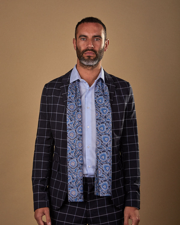 Three quarters view wearing 'The DeZon' blue paisley silk scarf draped around neck and paired with pale blue shirt and dark chequered suit.