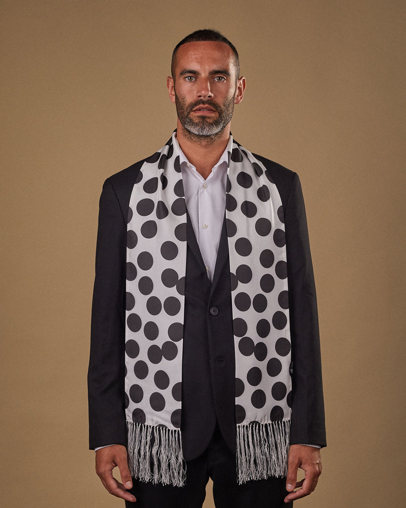 Portrait view of male model looking straight ahead, wearing 'The Gable' silk aviator scarf. Draped loosely around shoulders and neck and paired with a smart, dark suit.