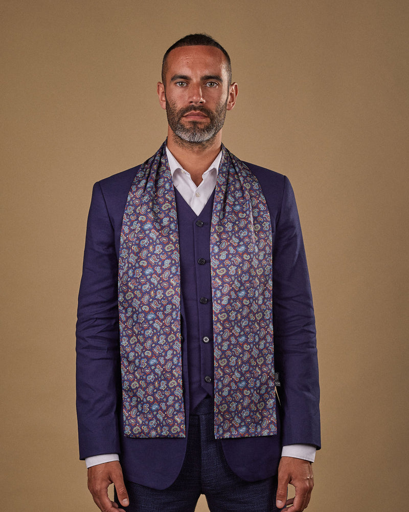Portrait view of male model looking straight ahead, wearing 'The Lexington' polyester scarf. Draped loosely around shoulders and neck and paired with a smart, midnight-blue suit.