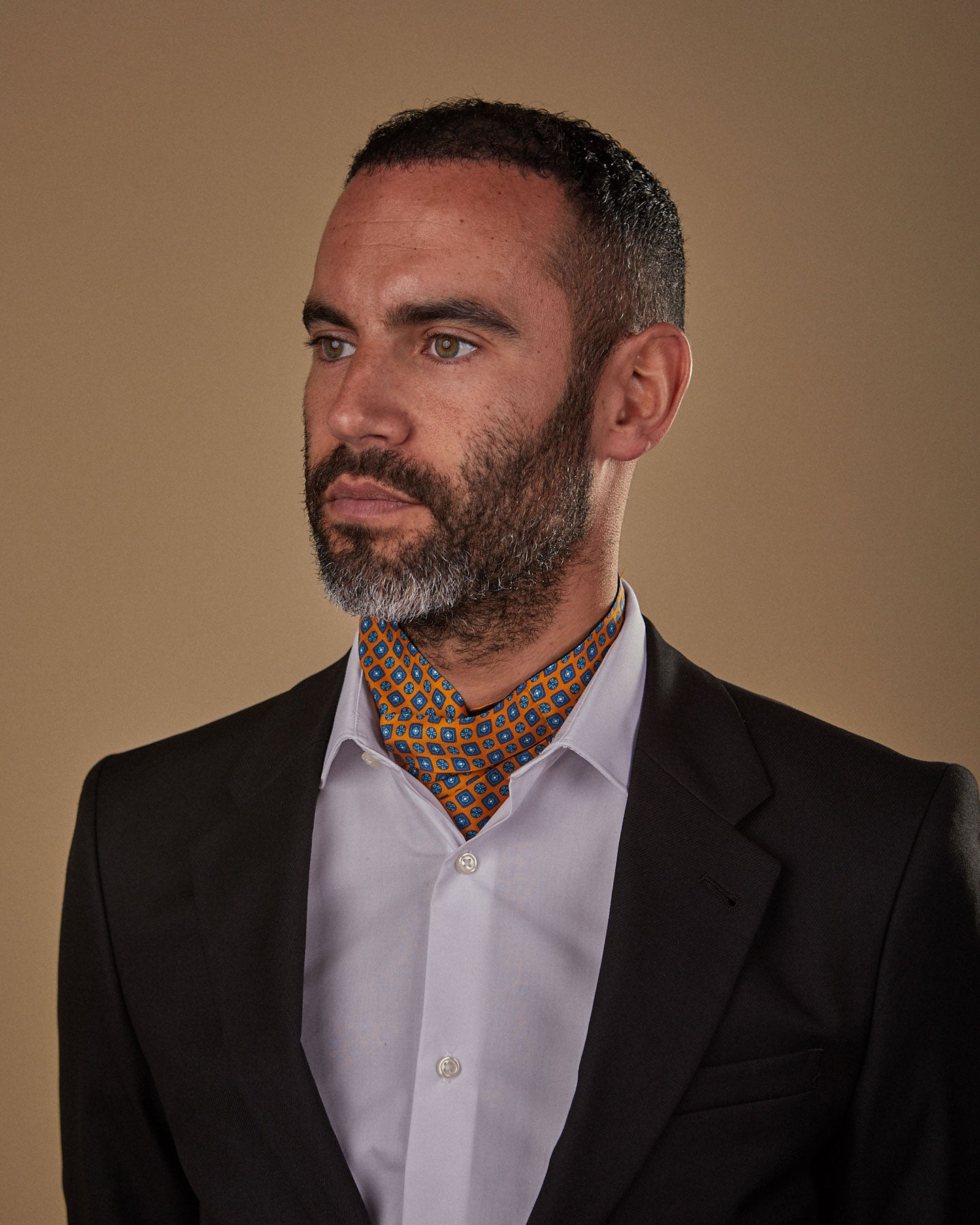 Model looking to his right, wearing gold and blue geometric patterned 'Madrid' single pointed Ascot tie. Neatly tucked under white shirt and paired with charcoal jacket.