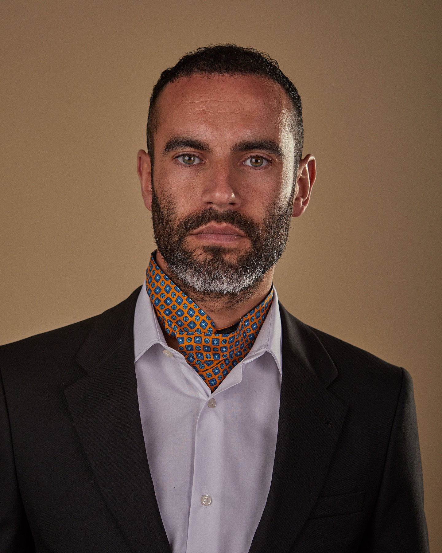 Model looking straight ahead wearing gold and blue 'Madrid' single pointed Ascot tie tucked neatly under white shirt. Paired with charcoal jacket.