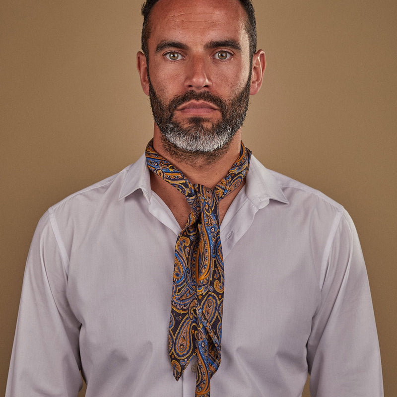 Man wearing 'The Ormond' multicoloured paisley and black bandana. Tied snugly around neck in a simple knot and paired with a white shirt.