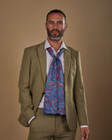 Facing ahead with hand in pocket, wearing a loosely tied Oxford polyester scarf in blue paisley with red ground. Paired with pale blue shirt and sandy-green suit.