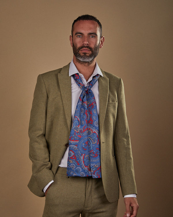 Front view with hand in pocket, wearing an Oxford silk scarf in blue paisley with red ground. Paired with pale blue shirt and sandy-green suit.