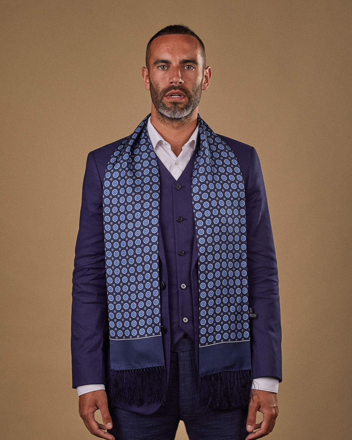 Portrait view of male model looking straight ahead, wearing 'The Palm' silk aviator scarf. Draped loosely around shoulders and neck and paired with a smart, midnight-blue suit.
