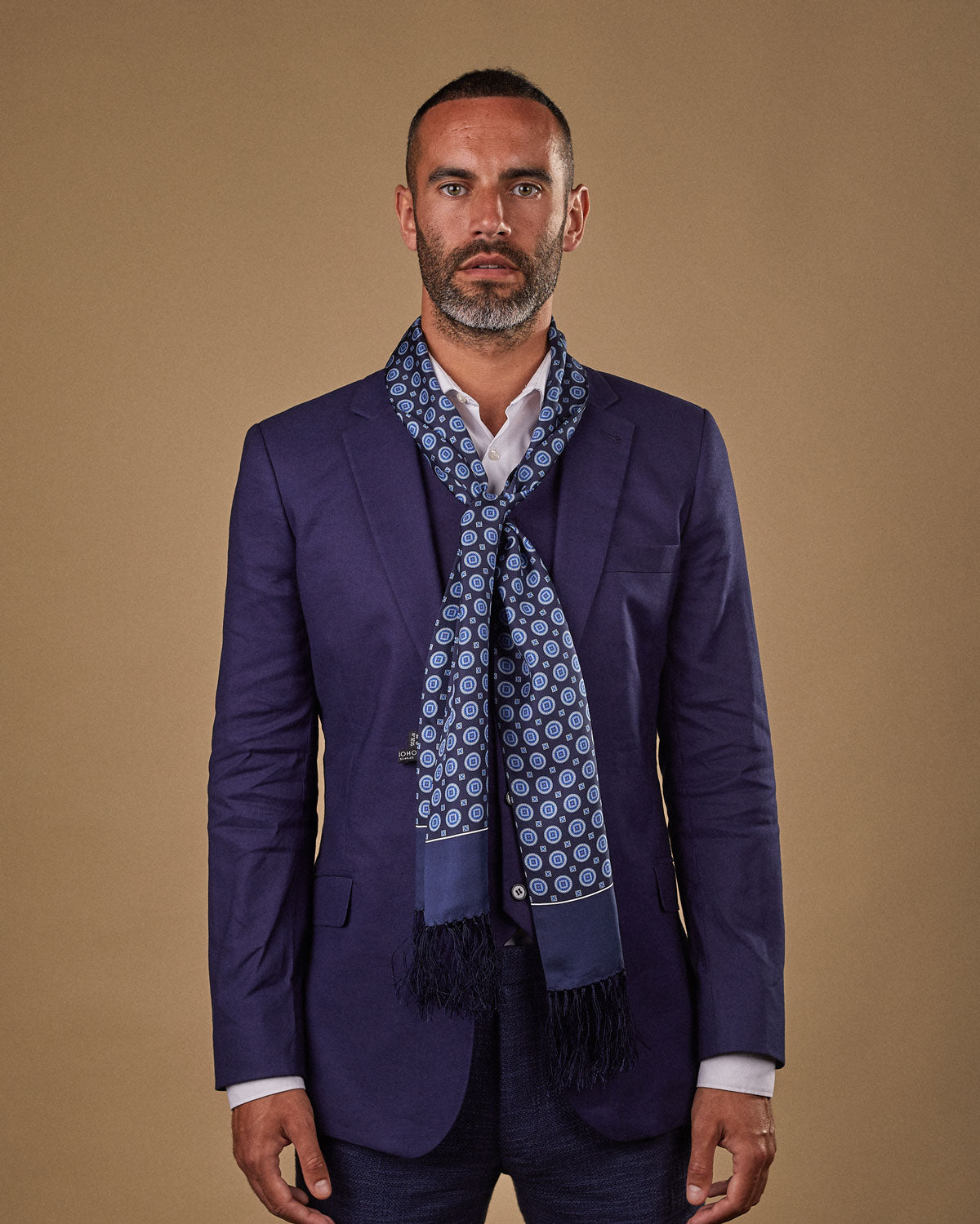 Portrait view of male model wearing 'The Palm' silk aviator scarf. Tied loosely around neck in a simple knot and shoulders and paired with a smart, midnight-blue suit.