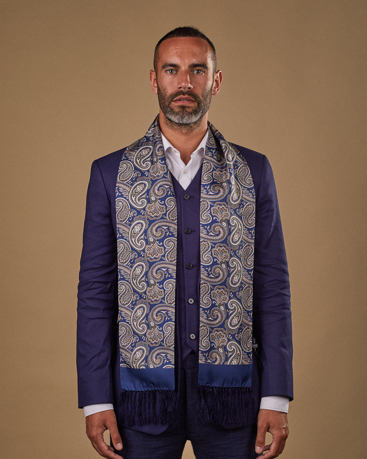 Portrait view of male model looking straight ahead, wearing 'The Redmond' silk aviator scarf. Draped loosely around shoulders and neck and paired with a smart, midnight-blue suit.
