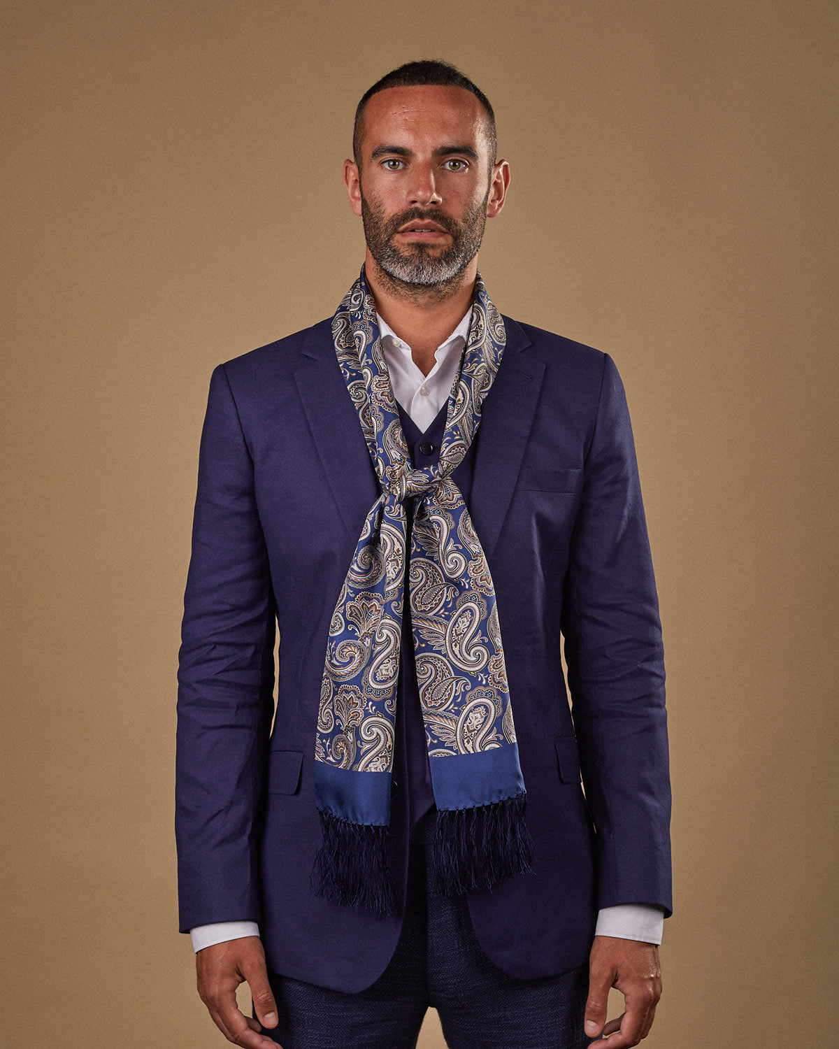 Portrait view of male model wearing 'The Redmond' silk aviator scarf. Tied loosely around neck in a simple knot and shoulders and paired with a smart, midnight-blue suit.