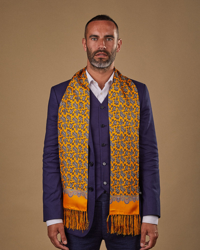 Portrait view of male model looking straight ahead, wearing 'The Richmond' silk aviator scarf. Draped loosely around shoulders and neck and paired with a smart, midnight-blue suit.