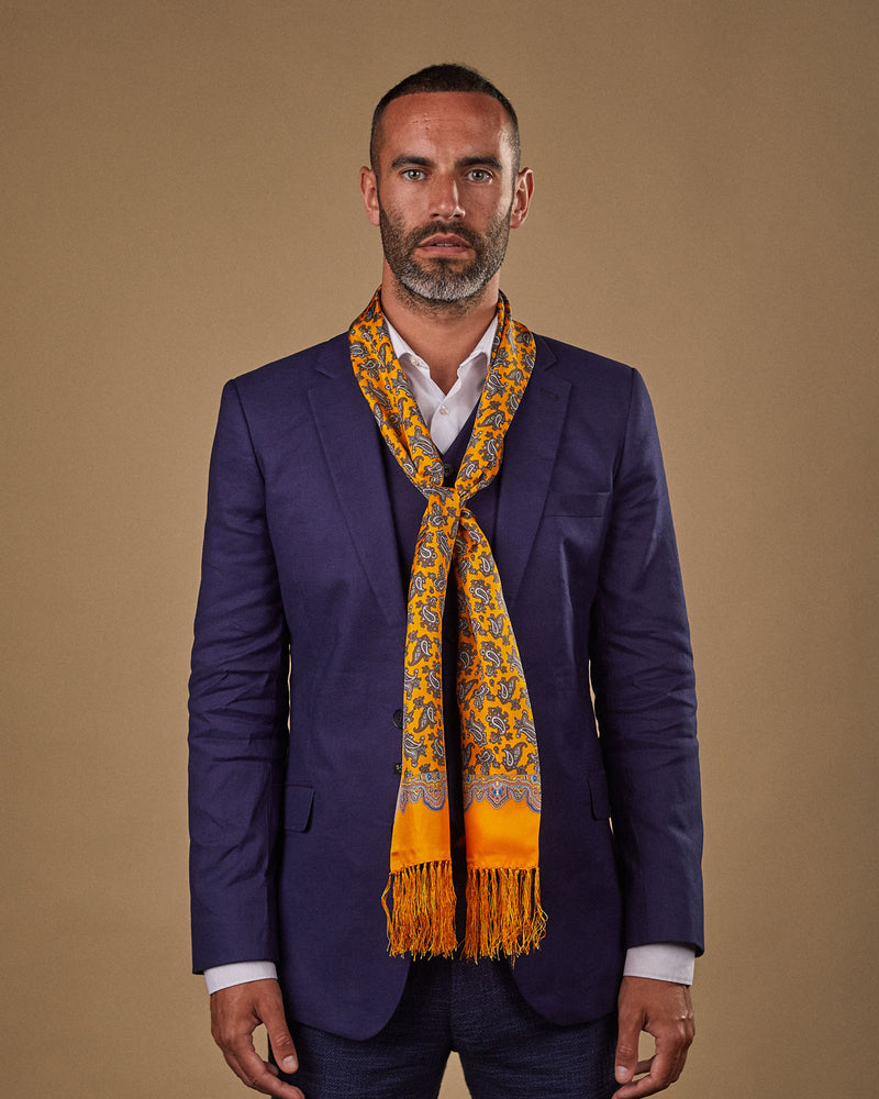 Portrait view of male model wearing 'The Richmond' silk aviator scarf. Tied loosely around neck in a simple knot and shoulders and paired with a smart, midnight-blue suit.