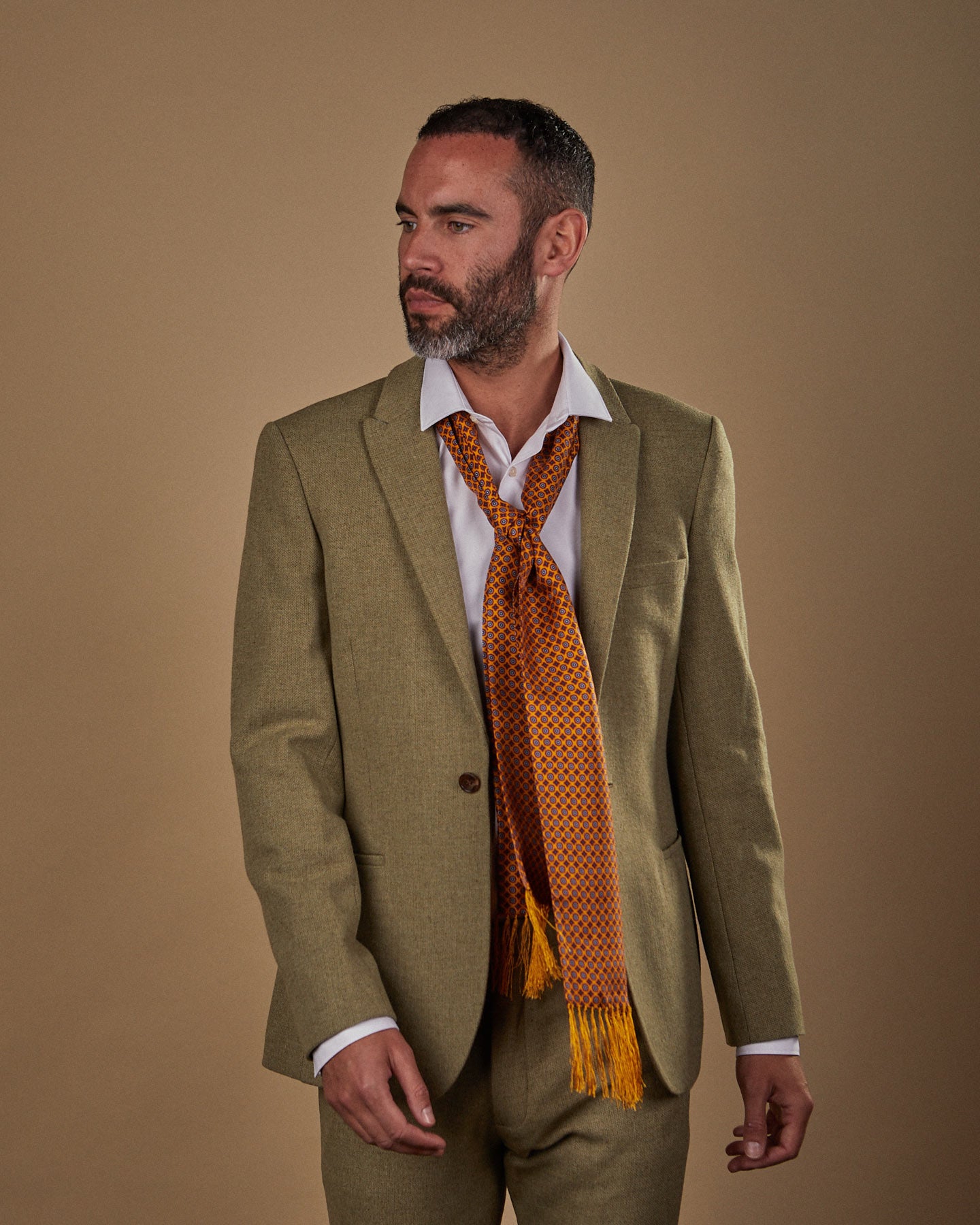 Facing right with hands by side, wearing the 'Toshima' silk aviator scarf in small geometric floral patterns with deep golden-orange ground. Paired with white shirt and sandy-green suit.