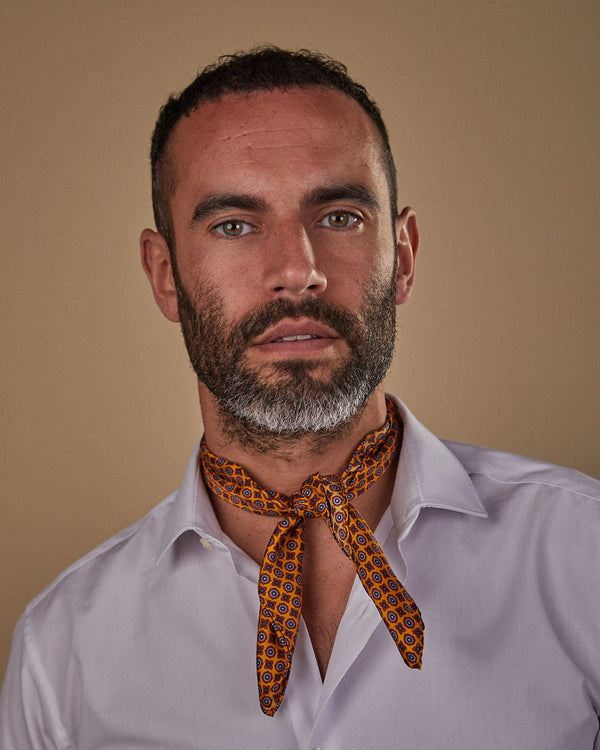Head and shoulders view of model facing straight ahead, wearing the 'Toshima' silk neckerchief with knot tie at front. Featuring small geometric floral patterns with deep golden-orange ground. 