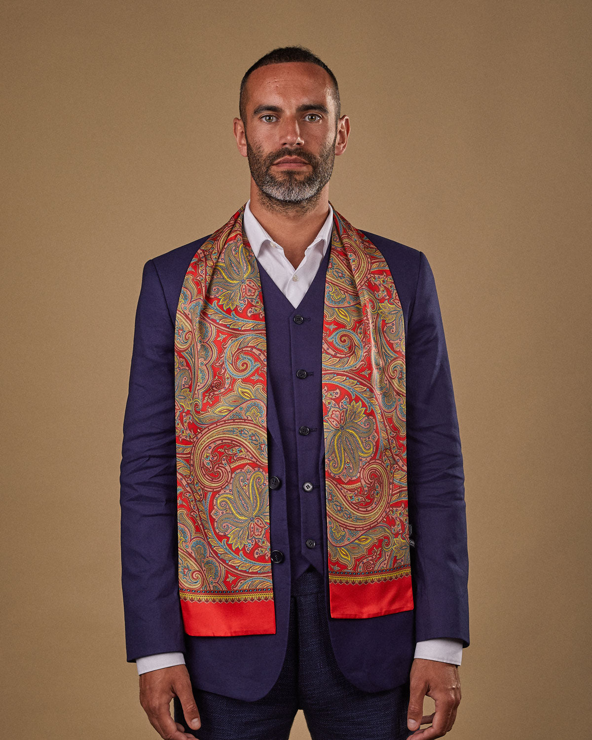 Portrait view of male model looking straight ahead, wearing 'The Victoria' silk scarf. Draped loosely around shoulders and neck and paired with a smart, midnight-blue suit.