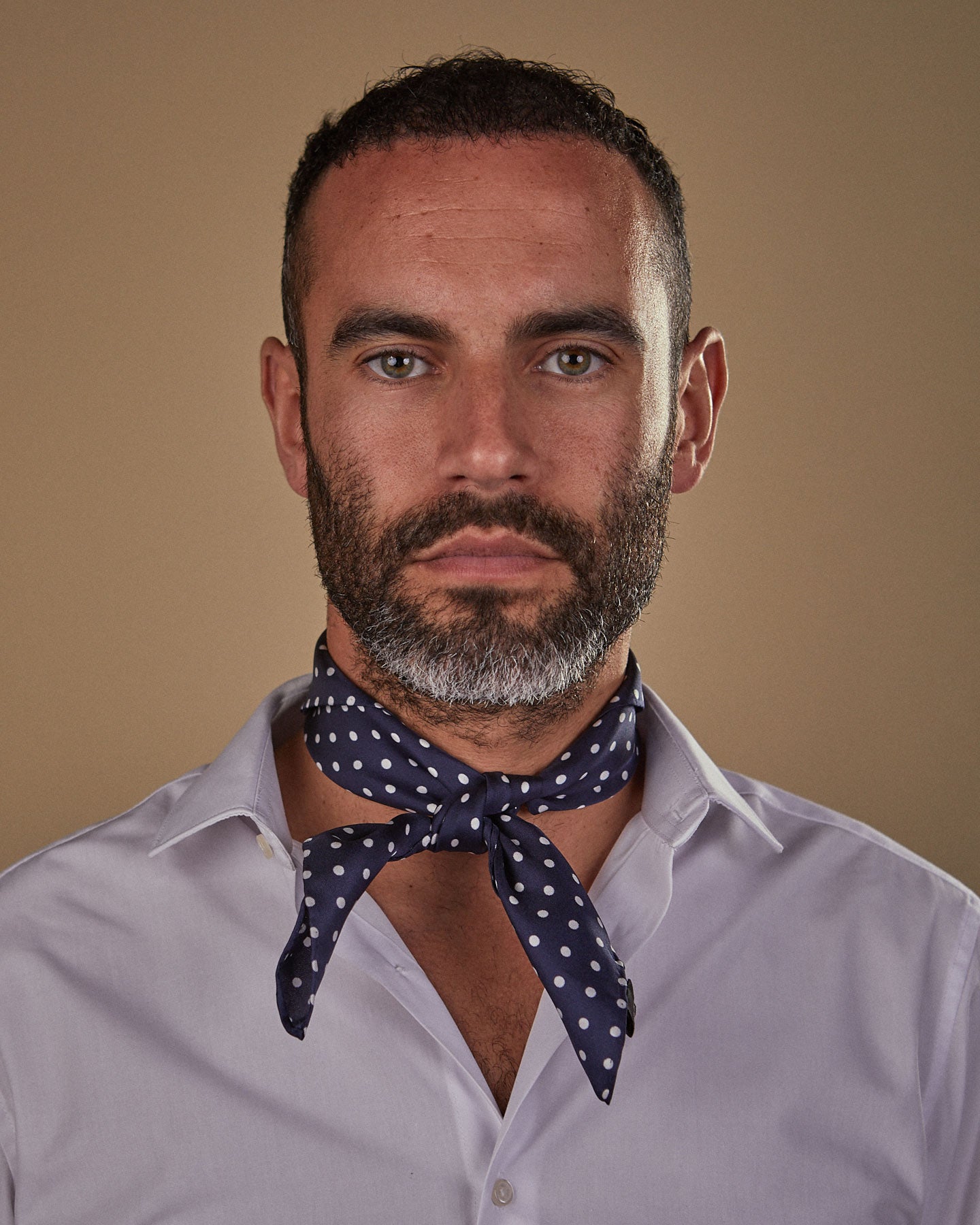 Head and shoulders view of model facing straight ahead, wearing the 'Westminster' silk neckerchief with knot tie at front. Featuring small white polka-dots on a dark blue ground. 