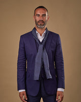 Portrait view of male model wearing 'The Westminster' silk scarf. Tied loosely around neck in a simple knot and shoulders and paired with a smart, midnight-blue suit.
