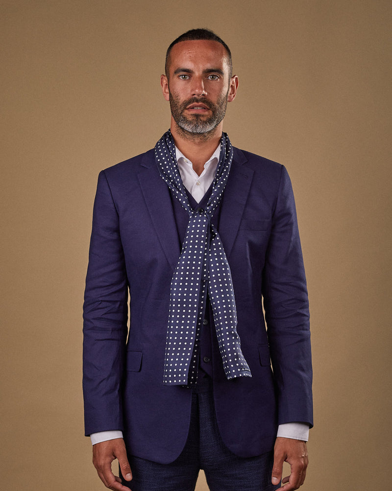 Portrait view of male model wearing 'The Westminster' silk scarf. Tied loosely around neck in a simple knot and shoulders and paired with a smart, midnight-blue suit.