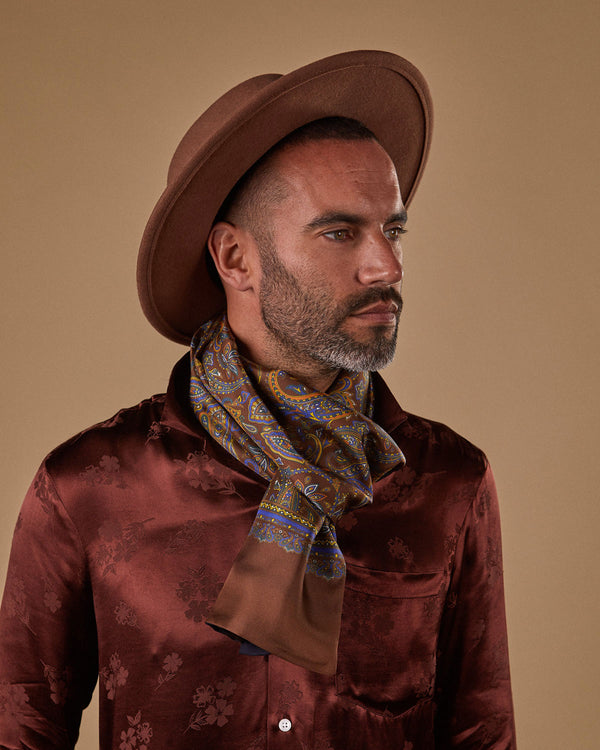 Model wearing 'The Whistler' wider multi-coloured paisley patterned wide dress scarf on a brown ground.