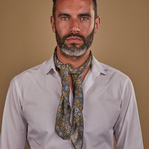 Man wearing 'The Whitehorse' grey and yellow multi-patterned bandana. Tied snugly around neck in a simple knot and paired with a white shirt.