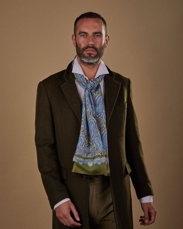 Front view wearing an olive green and blue paisley 'Abraham' silk wide dress scarf from SOHO Scarves, paired with white shirt, fawn trousers and long brown overcoat.