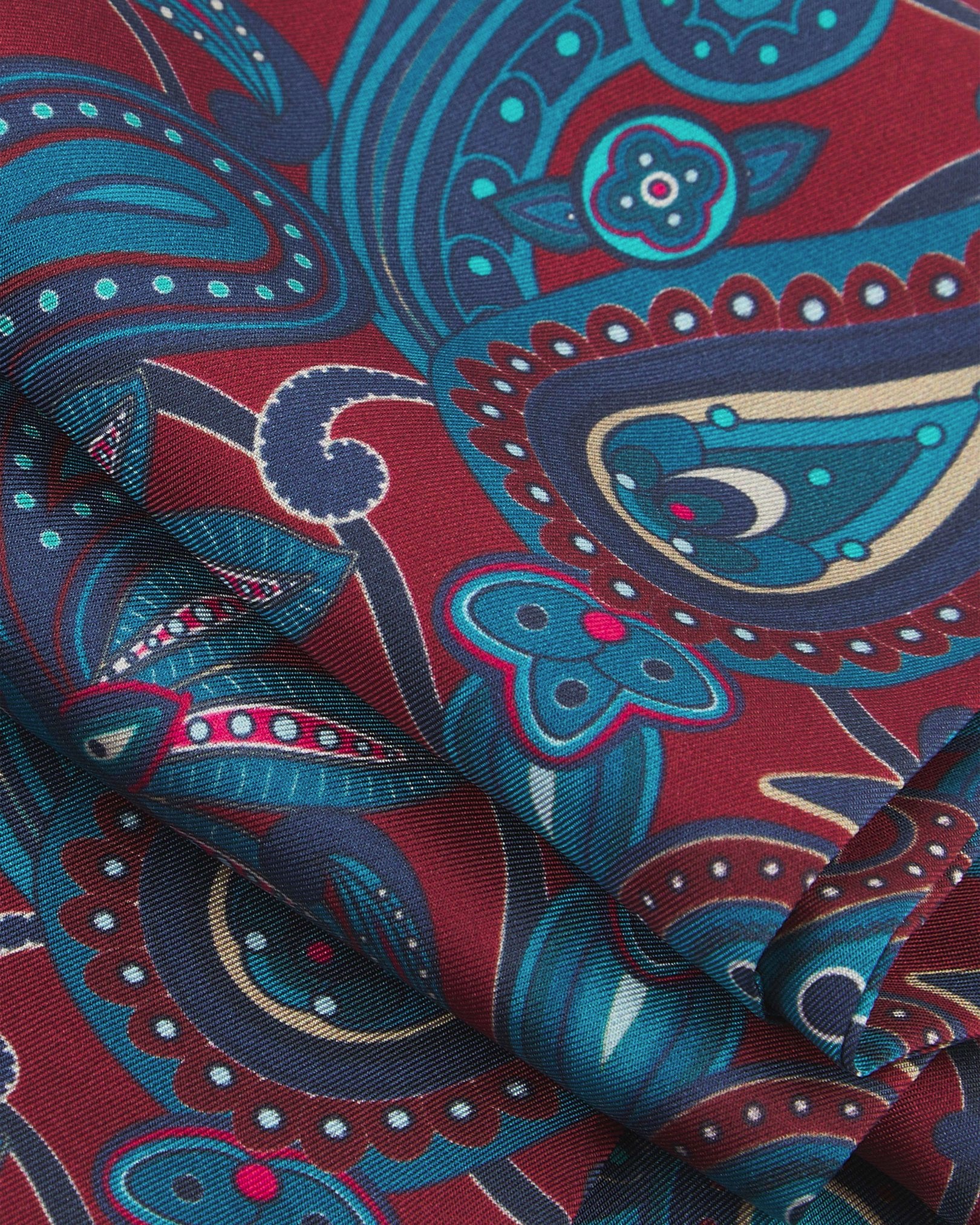 A ruffled close-up of the 'Windermere' rust silk scarf, presenting the overlapping elements of blue and blue-green paisley patterns.