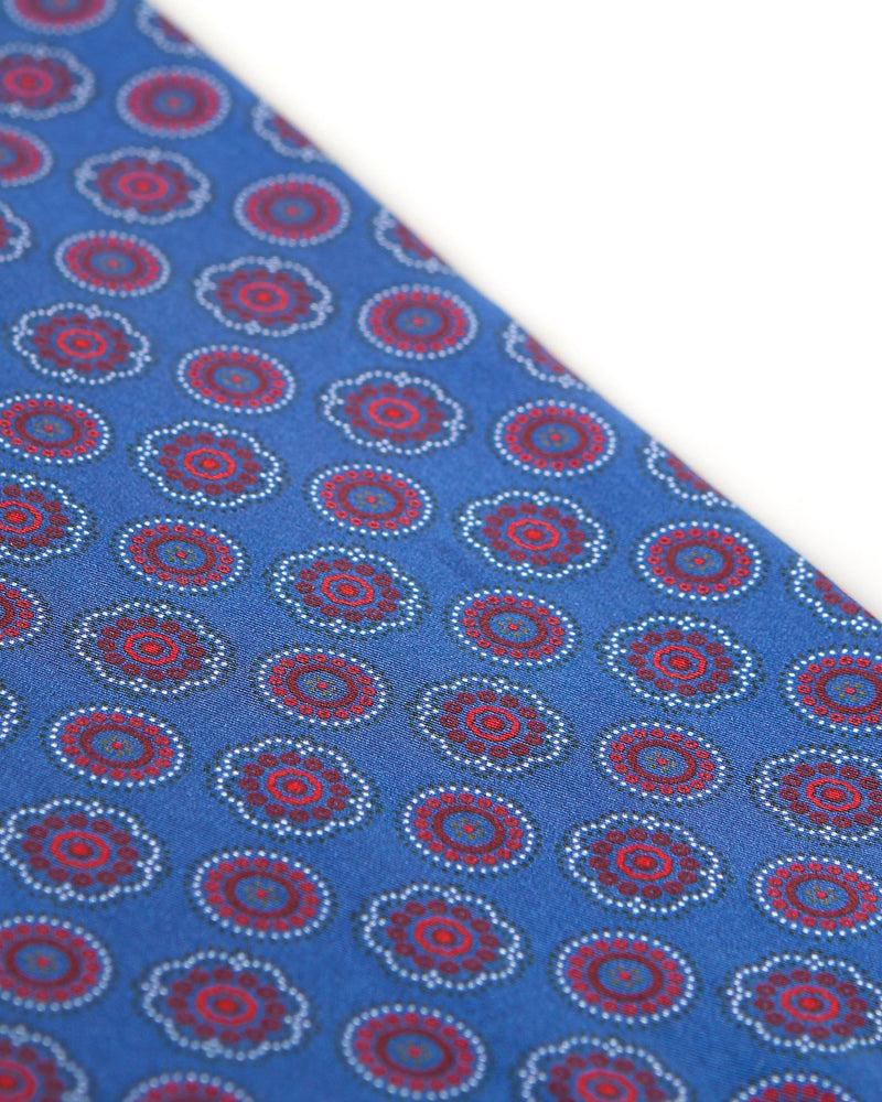 Angled view of the blue patterned 100 percent silk scarf, presenting a closer view of the small red and maroon circular and stylised floral patterns.