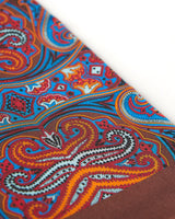 Angled closer view of the copper-brown silk scarf with focus on the large, ornate patterns in blue, orange, white and red.